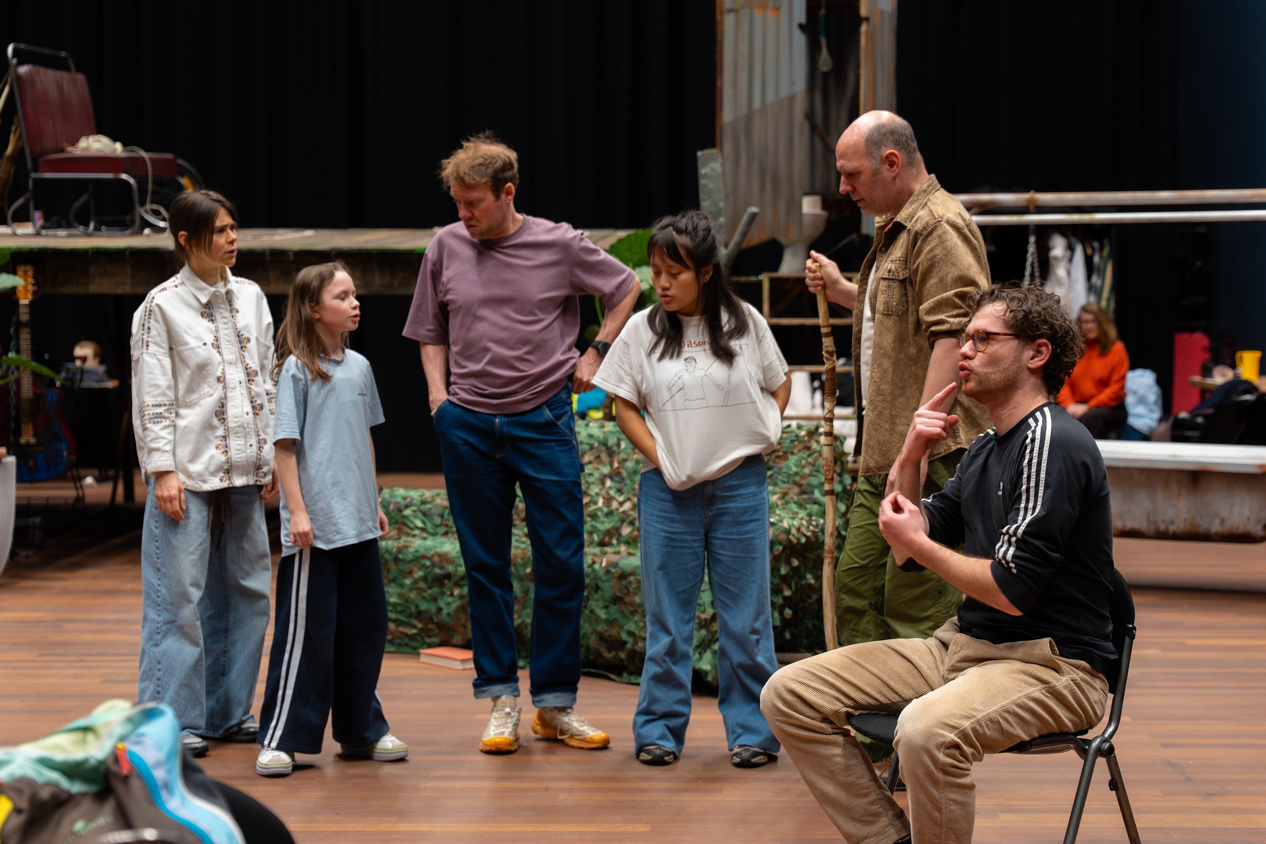Boaz gebaart zittend op een stoel wat de groep acteurs op de achtergrond zeggen.